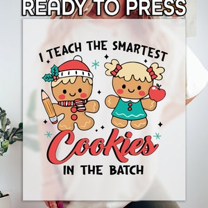 May include: White t-shirt with a red, white, and blue gingerbread person design. The text reads "I teach the smartest cookies in the batch".