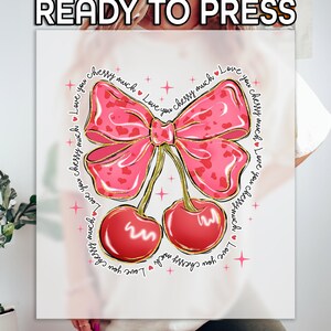 May include: A white t-shirt with a pink bow and two red cherries. The bow has a gold outline and a pink and white polka dot pattern. The cherries have gold outlines and are surrounded by small stars. The text "Love you cherry much" is repeated around the design.