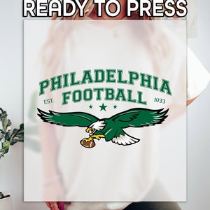 May include: White t-shirt with a green and white Philadelphia Football logo. The logo features an eagle with a football in its talons and the text "Philadelphia Football" with "Est. 1933".