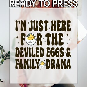 May include: White t-shirt with a brown graphic that says "I'm just here for the deviled eggs & family drama". The graphic includes a cartoon deviled egg.