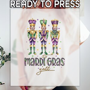 May include: A white t-shirt with a design of three purple and green Mardi Gras characters with gold accents. The text "it's Mardi Gras y'all" is written in gold below the characters.