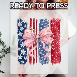 May include: A white graphic tee with a watercolor design featuring a pink bow adorned with flowers, set against a backdrop of red, white, and blue stripes and stars. The text "READY TO PRESS" is at the top.