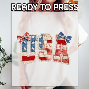 May include: A white t-shirt featuring the text USA in a patchwork style. The letters are decorated with red, white, and blue patterns, including stars and gingham checks. A red gingham bow sits on the letter U, and a blue bow sits on the letter A.