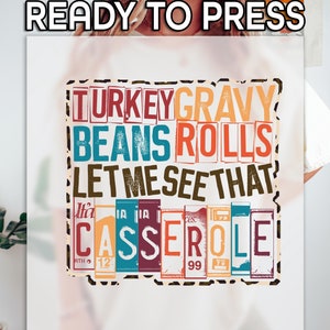 May include: A white ready-to-press sublimation transfer featuring the text "TURKEY GRAVY BEANS ROLLS LET ME SEE THAT CASSEROLE" in various colors. The design is framed with a black and brown border.