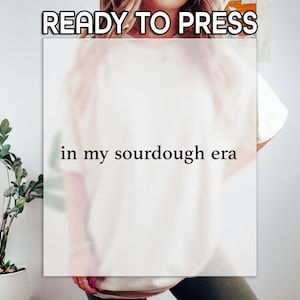 May include: A white t-shirt with the text "in my sourdough era" in a black font. The words "READY TO PRESS" are at the top of the image. The shirt is being worn by a person.