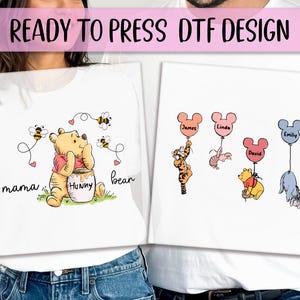 May include: Two white t-shirts with iron-on transfers. One shirt features Winnie the Pooh holding a honey pot with the text "mama bear". The other shirt features four Winnie the Pooh characters with Mickey Mouse ears and balloons with names "James", "Linda", "David", and "Emily".