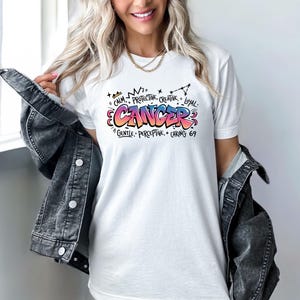 K&ouml;nnte beinhalten: Wei&szlig;es T-Shirt mit einem farbenfrohen Krebs-Grafik. Das Design enth&auml;lt das Wort "CANCER" im Graffiti-Stil, zusammen mit W&ouml;rtern wie "Ruhig", "Sch&uuml;tzend", "Kreativ" und "Loyal". Das Shirt hat auch die Nummer "69".