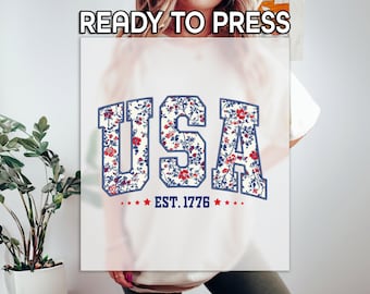 May include: White t-shirt with a blue and red floral print of the word "USA" and the text "EST. 1776" below.