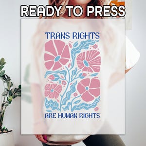 May include: White t-shirt with a graphic design of pink flowers and blue leaves. The text "Trans Rights Are Human Rights" is printed in blue below the flowers.