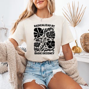 Może przedstawiać: Kremowy t-shirt z czarnym napisem "RADICALIZED BY BASIC DECENCY". Koszulka ma czarny kwiatowy wz&oacute;r. Modelka ma na sobie dżinsowe szorty i beżowy kardigan.