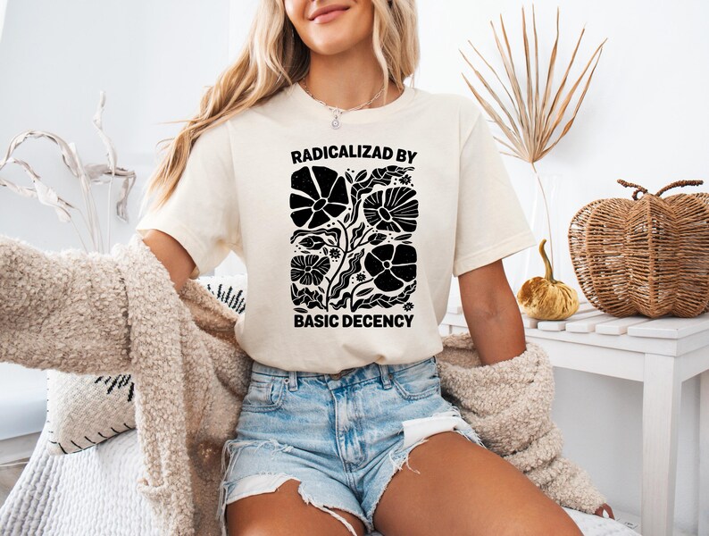 Może przedstawiać: Kremowy t-shirt z czarnym napisem "RADICALIZED BY BASIC DECENCY". Koszulka ma czarny kwiatowy wz&oacute;r. Modelka ma na sobie dżinsowe szorty i beżowy kardigan.