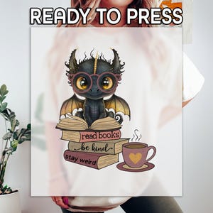 May include: A white t-shirt with a black dragon wearing glasses and sitting on a stack of books. The books have the text "read books", "be kind", and "stay weird". There is a cup of coffee next to the dragon.