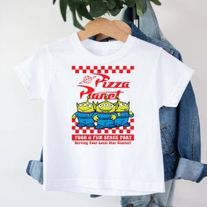 May include: White t-shirt with a red and white checkered design and the words "Pizza Planet" in red. Three green aliens from the Toy Story movie are standing in front of the text. The text "Food & Fun Space Port" and "Serving Your Local Star Cluster!" are printed below the aliens.