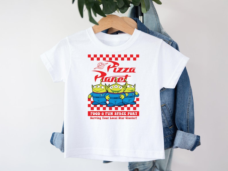 May include: White t-shirt with a red and white checkered design and the words "Pizza Planet" in red. Three green aliens from the Toy Story movie are standing in front of the text. The text "Food & Fun Space Port" and "Serving Your Local Star Cluster!" are printed below the aliens.