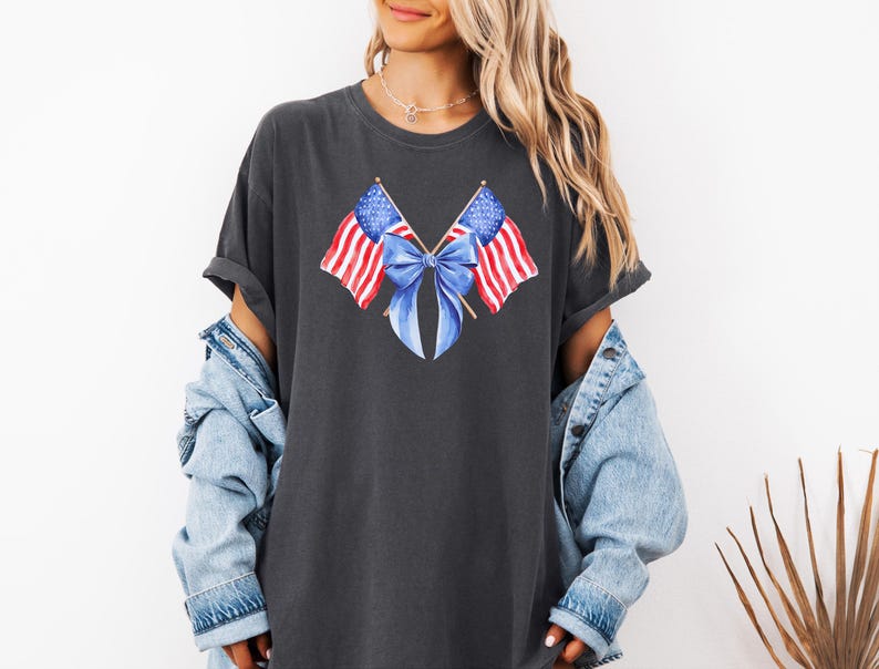 Puede incluir: Camiseta gris oscuro con un dise&ntilde;o de acuarela de dos banderas americanas cruzadas con un lazo azul. Las banderas tienen rayas rojas y blancas y estrellas azules. La camiseta se usa con una chaqueta vaquera.