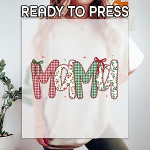 May include: A white graphic featuring the word "MAMA" in a festive design. The letters are decorated with red gingham, green plaid, and floral patterns, accented with bows and stars. The text "READY TO PRESS" is at the top.