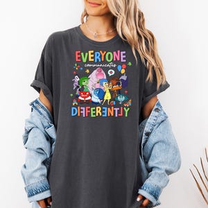 May include: A dark gray t-shirt with a colorful graphic of characters from the movie Inside Out. The text "Everyone communicates differently" is printed in rainbow letters on the shirt.