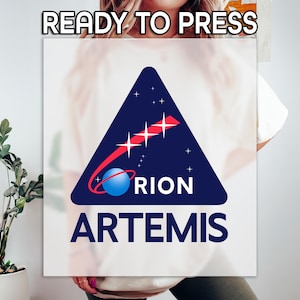 May include: A white rectangular graphic with the text "READY TO PRESS" at the top. The graphic features a navy blue triangle with the words "ORION" and "ARTEMIS" in white. A red comet-like design and a blue planet are also present.