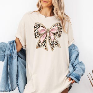 May include: A beige t-shirt with a large leopard print bow with pink ribbon accents. The bow has gold glitter accents.
