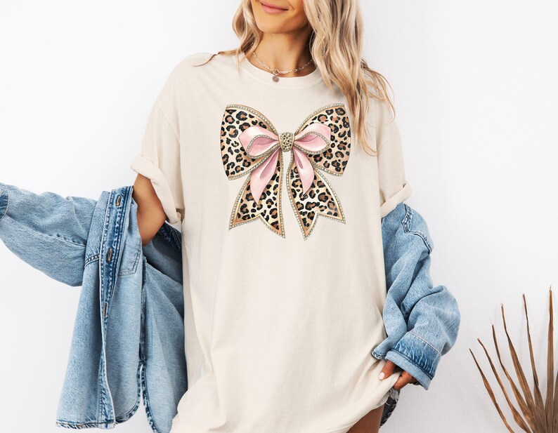 May include: A beige t-shirt with a large leopard print bow with pink ribbon accents. The bow has gold glitter accents.