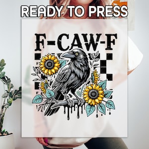 May include: A white graphic tee with a black raven illustration perched on a branch with sunflowers and the text "F-CAW-F". The design includes a black and white checkered pattern and the words "READY TO PRESS" at the top.