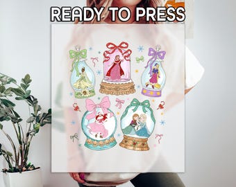 Könnte beinhalten: Weißes T-Shirt mit einer farbenfrohen Grafik von sechs Schneekugeln, die jeweils eine andere Prinzessin zeigen. Die Schneekugeln sind mit Schleifen und Zuckerstangen verziert. Der Text oben lautet "READY TO PRESS".