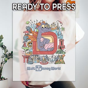 May include: White t-shirt with a retro design featuring a purple dragon inside a red letter "D". The design includes various Disney World landmarks and the text "Walt Disney World" below. The words "READY TO PRESS" are at the top.
