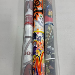 May include: Three rolls of wrapping paper with various designs. The first roll has a red and white design with the text "Santa's Workshop". The second roll has a colorful design with the text "Pop-Up's". The third roll has a black and white design with the text "1970's Summer Jams".