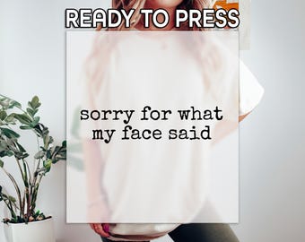 May include: Cream-colored t-shirt with the text "sorry for what my face said" in a bold, black font. The words "READY TO PRESS" are at the top. The shirt is displayed on a person.