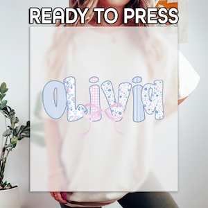 May include: A white t-shirt with the name "Olivia" in a playful font. The letters feature various patterns, including blue stripes, floral prints, and pink gingham. A pink bow is placed below the name. The text "READY TO PRESS" is at the top.