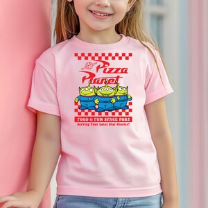 May include: Pink t-shirt with a graphic of three green aliens from the movie Toy Story. The aliens are standing in front of a red and white checkered sign that says "Pizza Planet" and "Food & Fun Space Port".