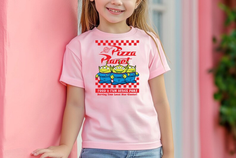 May include: Pink t-shirt with a graphic of three green aliens from the movie Toy Story. The aliens are standing in front of a red and white checkered sign that says "Pizza Planet" and "Food & Fun Space Port".