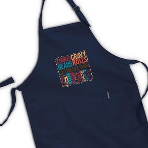 May include: Navy blue apron with two front pockets and adjustable neck strap. The apron features a colorful text design that reads "TURKEY GRAVY BEANS ROLLS LET ME SEE THAT CASSEROLE".