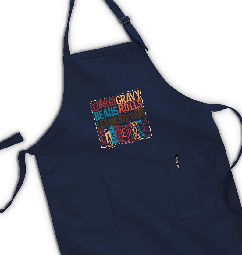 May include: Navy blue apron with two front pockets and adjustable neck strap. The apron features a colorful text design that reads "TURKEY GRAVY BEANS ROLLS LET ME SEE THAT CASSEROLE".