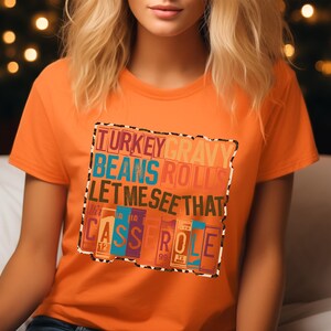 May include: Orange t-shirt with a colorful graphic that reads "TURKEY GRAVY BEANS ROLLS LET ME SEE THAT CASSEROLE". The text is in various colors and fonts, with a leopard print border. The shirt is short-sleeved.