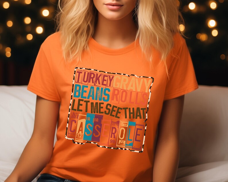 May include: Orange t-shirt with a colorful graphic that reads "TURKEY GRAVY BEANS ROLLS LET ME SEE THAT CASSEROLE". The text is in various colors and fonts, with a leopard print border. The shirt is short-sleeved.