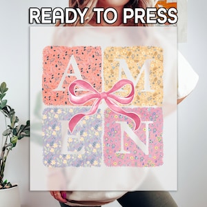 May include: White t-shirt with a floral design spelling out the word "AMEN" in white letters. A pink bow is centered on the design.