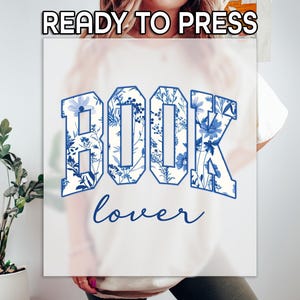 May include: White t-shirt with the words "BOOK lover" in blue. The word "BOOK" is in a collegiate style font, filled with a blue floral pattern. The text "READY TO PRESS" is at the top.