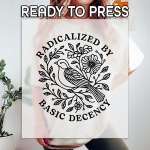 May include: A white t-shirt with a black graphic design. The design features a bird surrounded by leaves and flowers, with the text "RADICALIZED BY BASIC DECENCY" in a circular arrangement. The words "READY TO PRESS" are at the top.
