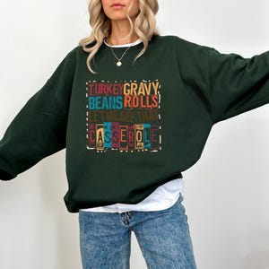 May include: A dark green sweatshirt with the text "TURKEY GRAVY BEANS ROLLS! LET ME SEE THAT CASSEROLE" in colorful block letters. The sweatshirt is a crew neck style and is worn with blue jeans.