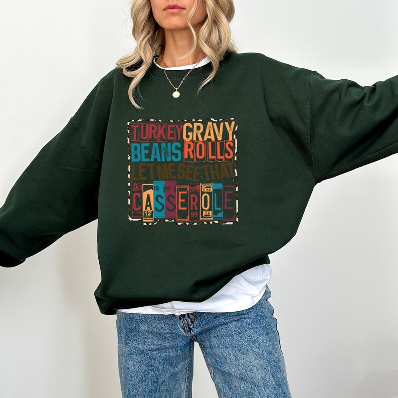 May include: A dark green sweatshirt with the text "TURKEY GRAVY BEANS ROLLS! LET ME SEE THAT CASSEROLE" in colorful block letters. The sweatshirt is a crew neck style and is worn with blue jeans.