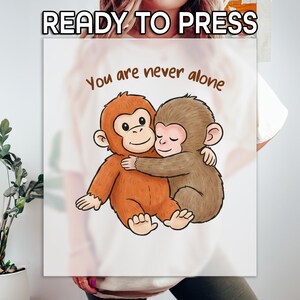 May include: A white sublimation print featuring two cartoon monkeys hugging. The text "You are never alone" is above the monkeys, and "READY TO PRESS" is at the top. The monkeys are brown and orange.