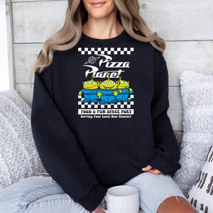May include: Black sweatshirt with a white checkered pattern and a graphic of three green alien characters from the movie Toy Story. The graphic says "Pizza Planet" and "Food & Fun Space Port" with the tagline "Serving Your Local Star Cluster!"