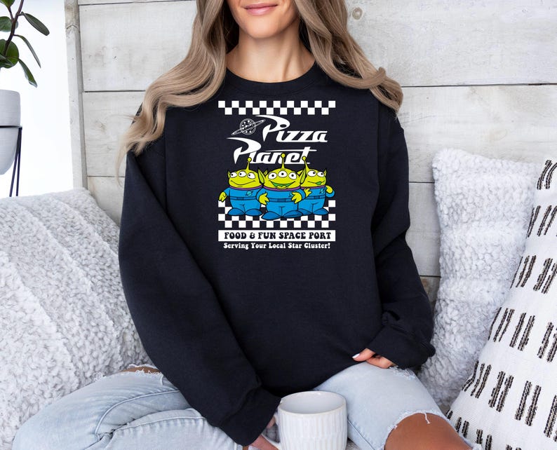 May include: Black sweatshirt with a white checkered pattern and a graphic of three green alien characters from the movie Toy Story. The graphic says "Pizza Planet" and "Food & Fun Space Port" with the tagline "Serving Your Local Star Cluster!"