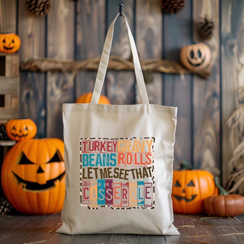 May include: A cream-colored tote bag with the words "TURKEY GRAVY BEANS ROLLS LET ME SEE THAT CASSEROLE" printed in colorful block letters. The bag has two handles and is hanging in front of a rustic wooden background with pumpkins.
