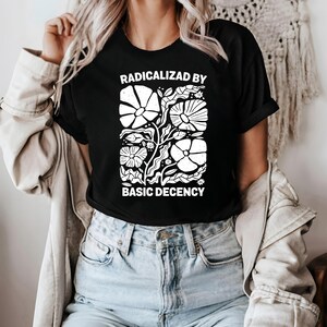 Może przedstawiać: Czarny t-shirt z białym kwiatowym wzorem i napisem "RADICALIZED BY BASIC DECENCY". Grafika przedstawia stylizowane kwiaty i liście. Odzież codzienna.