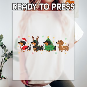 May include: A white t-shirt with the text "READY TO PRESS" at the top. Below the text are four cartoon dachshunds in festive holiday attire. The dogs are wearing a Santa suit, reindeer antlers, a Christmas tree costume, and a gingerbread design.