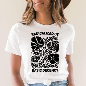 Może przedstawiać: Biała koszulka z czarnym kwiatowym wzorem i napisem "RADICALIZED BY BASIC DECENCY". Grafika zawiera stylizowane kwiaty i liście. Koszulka ma okrągły dekolt i kr&oacute;tkie rękawy.