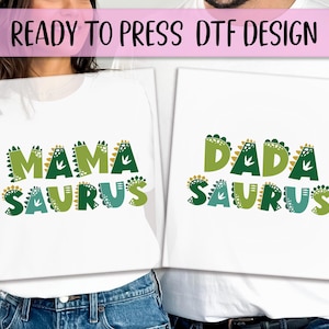 May include: White t-shirts with the phrases "MAMA SAURUS" and "DADA SAURUS" in green, teal, and gold dinosaur-themed lettering. The text "READY TO PRESS DTF DESIGN" is above the shirts in a pink banner.