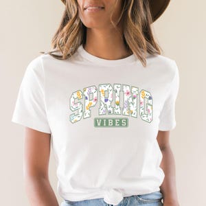 May include: White t-shirt featuring the text "SPRING VIBES" in a floral design. The text is in a collegiate-style font, with each letter filled with colorful flowers and leaves. A green rectangle below the text reads "VIBES".